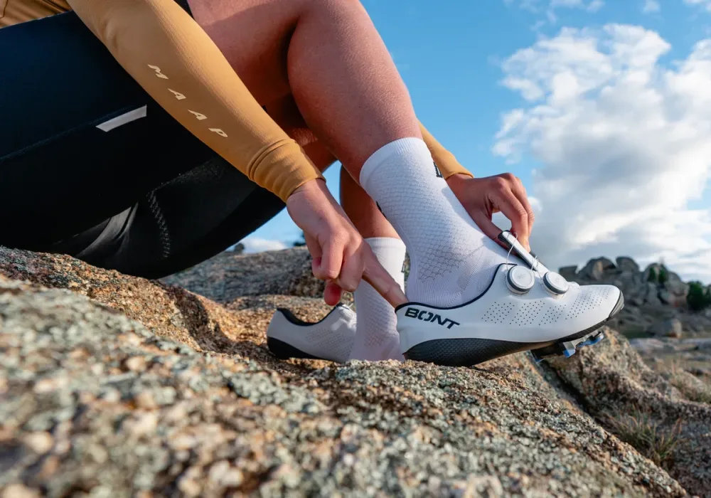 A rider fastening the dual BOA dials on a white Bont Riot 24 road shoe, sitting on a granite outcrop.