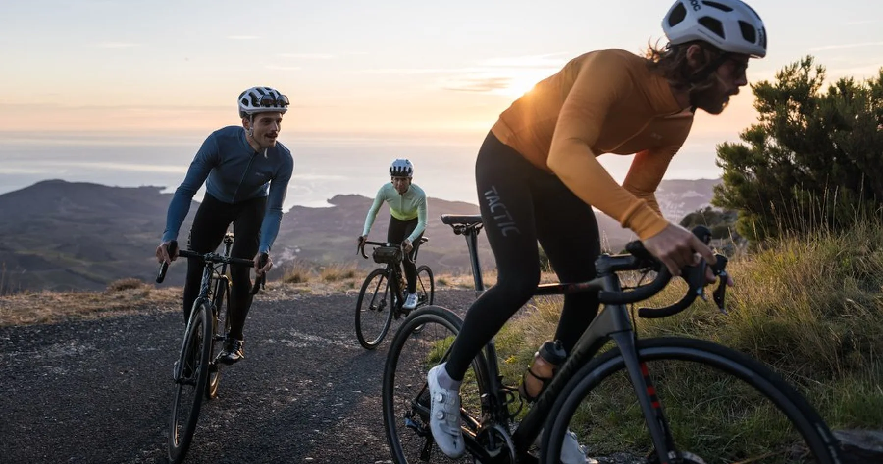 Three riders in Tactic kit climbing a coastal road at sunset